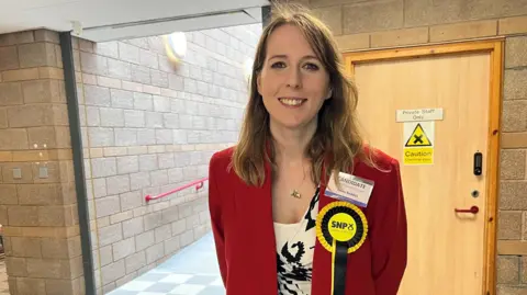 Emma Roddick is standing indoors. She has long fair hair and is wearing a red suit jacket over a black and white top. She is wearing a yellow and black rosette.