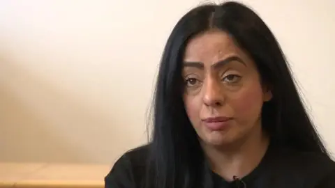 Oldham Council leader Arooj Shah looks forward. She has black shoulder length hair and is wearing a black top. She is wearing a lapel mic for an interview. 