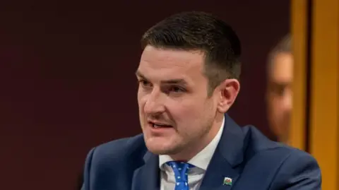 Getty Images A man with short brown hair and a blue suit and blue tie with a small Welsh national flag badge on the lapel, James Evans.