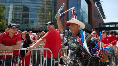 EPA People wearing red clothing lean against security railing while a man in a cowboy hat hawks souvenirs
