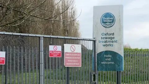 The entrance to the Oxford sewage treatment works with Thames Water sign that says "Hello and welcome", with other signs including No Parking and Gates in constant use 