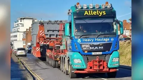 A large lorry seen from the front which has a blue and turquoise cab and at the rear you can see large grey industrial objects with buildings and cars seen in the background.