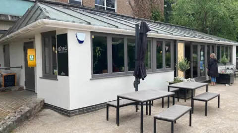 The picture shows the outdoor of The Shed. The walls are white and there are grey frames around the windows. There are benches and chairs outside.