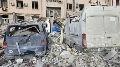 Karol Swiacki A close up of the damage to Karol's van. WIth the beige building hit by the missile in the background. All the glass from the windows is gone, from the building and the vans, they are covered and surrounded by rubble.