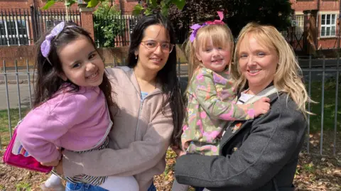 Two mums and their daughters are smiling for the photo in a park near the Saffron pre-school in Leicester. 