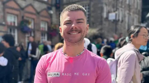 Chris Hall. Chris is a white man in his 20s with cropped brown hair and a stubbled beard. She smiled for the camera as she stood on a busy street in Edinburgh for the Fringe Festival. She was wearing a bright pink shirt and a beaded necklace. 