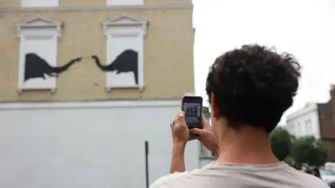 PA Media Un hombre toma una fotografía de una obra de Banksy con dos elefantes en su teléfono