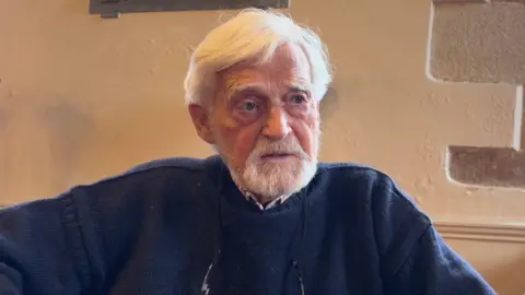 A man sits in front of a white stone wall. He has a white beard and white hair. He is wearing a navy blue jumper with a shirt underneath.