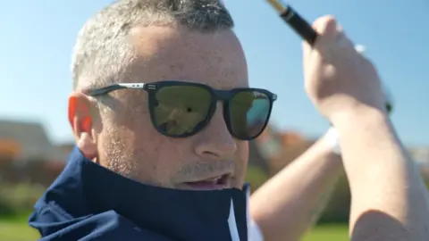 Jamie Niblock/BBC Paul Innocent has cropped grey hair and is wearing sunglasses and a navy jacket pulled up around his face. He is swinging a golf club.