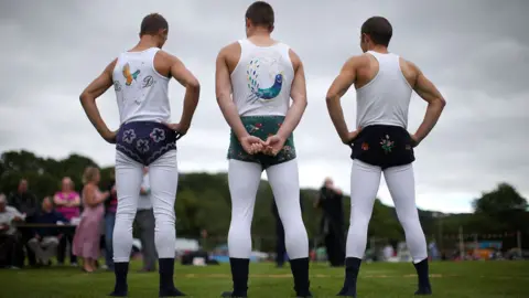 Getty Images Three men standing facing away. Two have hands on hips and one has hands behind his back. They are all wearing white long johns and white vest tops. The tops of two have birds embroidered on them - one is a hummingbird and one is peacock. All three are wearing velvet trunks - blue with embroidered flowers, turquoise with an unclear pattern, and black with embroidered flowers. In front of them, and slightly out of focus, is a grassy field, trees, fells and some spectators. 