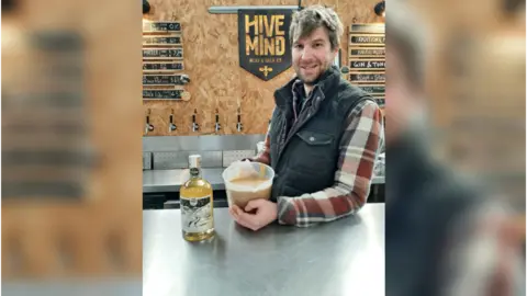 Wye Valley Meadery Matt smiling to camera behind a stainless steel countertop in his meadery with a bottle of his mead and a measuring jug containing the waste yeast.