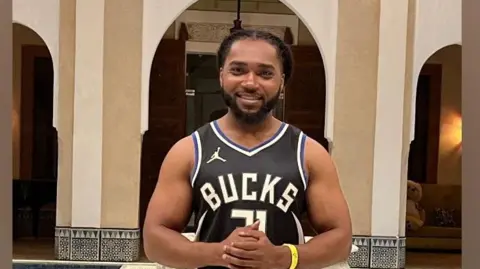 Person in a black Milwaukee Bucks  jersey and shorts standing indoors before ornate tiled arches and a decorative fountain 