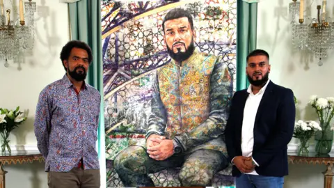 BBC Jack, left, and Rizwan stand beside a vibrant, mosaic-inspired portrait of Rizwan in an elegant room with chandeliers and floral arrangements.