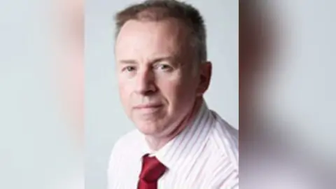 Warwickshire County Council A short-haired man with a pink and white stripy shirt and red tie looks at the camera.
