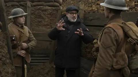 Entertainment One Director Sam Mendes is dressed in black, standing in a trench, and explaining something to two actors dressed as Great War soldiers