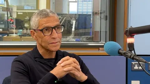 BBC Chris Hughton wearing glasses, sat in BBC Radio Surrey's studio. 