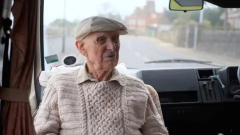 Bill wearing a cream cap and a beige jumper and shirt sat inside a van with the road behind him