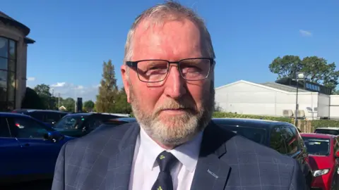 Mr Beattie has grey hair and a beard. He is wearing black square glasses, a navy checked blazer, a white shirt and a navy tie with green shamrocks on it. He is standing in front of a number of cars and a grey building. 