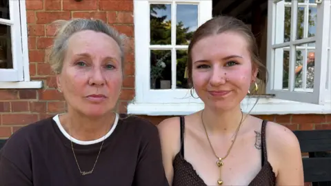 BBC/Richard Knights Two women are sitting outside in a pub garden, both looking at the camera. The one on the left is older than the one on the right.
