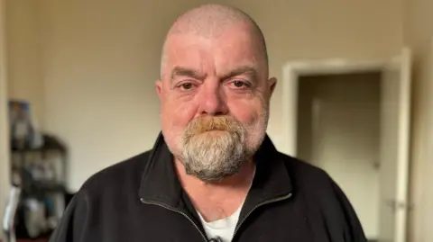 Paul Cowley has a goatee beard and grey hair cut very short. He is wearing a dark sweatshirt with small collar and zip, with a white t-shirt underneath. He is standing in a room with cream walls, with a blurred out bookshelf to the left and open door to the right.