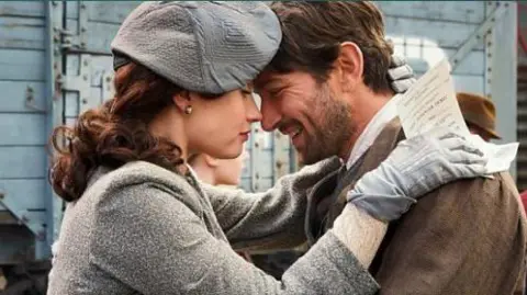 BBC Jessica Brown Findlay and Michiel Huisman pressing their foreheads together and smiling at one another. The woman is holding a letter in her hand on his shoulder. The picture is from the The Guernsey Literary and Potato Peel Pie Society film.