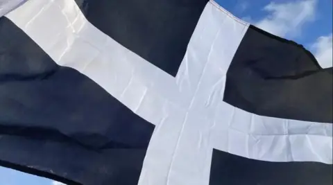 An unfurled black flag with a white cross and glimpses of the sky behind.