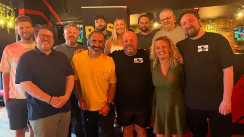 A group of eleven people made of 2 women and 9 men standing together smiling at the camera.