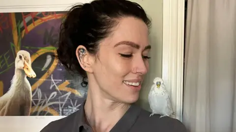 A woman with long dark hair pulled back behind her head looks to her left shoulder when a white budgie with a yellow beak is perched.