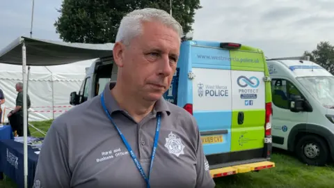 Graham Donaldson wears a grey tshirt with a blue lanyard around his neck. He has grey hair. He i standing on grass in front of a blue and green West Mercia Police van.