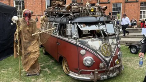Becki Bowden/BBC A person is dressed in a long brown outfit with a red alien type mask, holding a skull on a stick and stood next to a brown campervan with metal objects stuck on the top. 