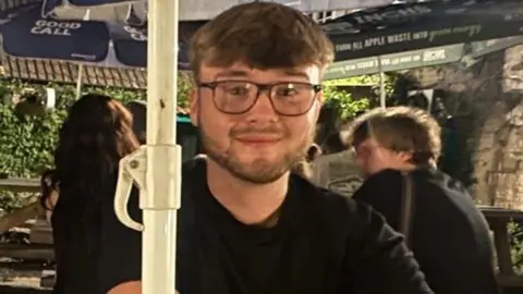 Ayden Strawson A young man with brown hair and glasses sitting at an outdoor table in a beer garden with other people in the background. 