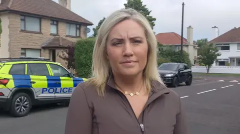 Diane Forsythe has shoulder length blonde hair and is wearing makeup, a gold necklace and a brown zip-up top. She is standing in front of a house with a yellow, blue and grey police jeep behind her.