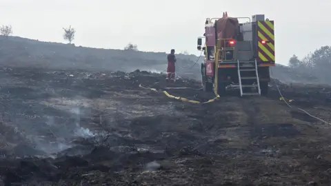 Jason Darkin A firefighter vehicle - not a typical fire engine but a vehicle adapted for more challenging terrain - parked in the middle of a scorched landscape. Steam rises where fires have recently been extinguished. A lone firefighter stands in the centre, surrounded by the coils of the yellow water pipes.