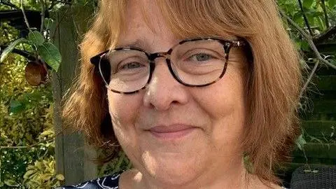 Kate Morris A woman wearing glasses poses outdoors in a garden, with green leaves and a wooden fence in the background.