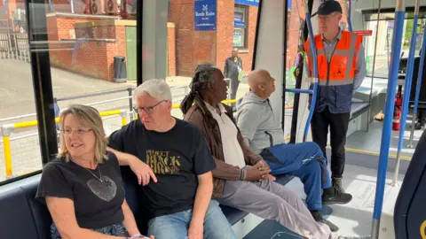 Passengers on the first public trial of Coventry's Very Light Rail system. They include a white man and a woman, both in black t-shirts, a black man with a brown jacket, sitting next to a bald man in a grey top, both looking at a council official in an orange hi-viz jacket, standing by the car's door.