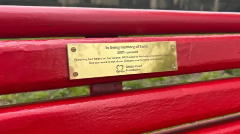 BBC/Chloe Hughes A close up of the red bench with grass beyond it. There is a gold plaque that reads "in living memory of Faith, 2001 to present. Wearing her heart on her sleeve, all thanks to the help of scientific research. But our work is not done. Donate now to keep it beating." underneath the text is the British Heart Foundation logo which is a heart.