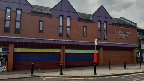 View of a two-storey retail unit in red brick, with high narrow windows on the first floor. The three units on the ground floor have roller shutters over then, and a sign on the building reads: Houghton-le-Spring Library and Learning Centre.