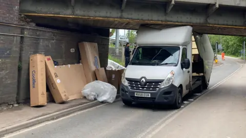 Katy Prickett/BBC A white van has crashed into a low railway bridge. There are several boxes on the side of the road leaning against part of the bridge.