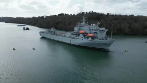 RAF Angus, a large navy vessel, in the middle of a river. The river water is blue. There are trees in the background. The sky is blue with white clouds. 