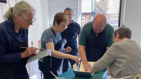 Dacorum Heritage Trust A group of volunteers holding pieces of paper, and all wearing aprons. They are all standing up, in a room, with a large window behind them. There are three women and two men. A building is in the distance. 