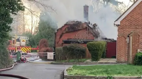 Smoke rises from the flattened thatched roof of the property which has a fire engine next to it.