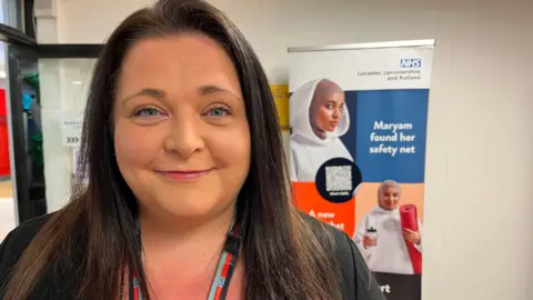 Lucy looks down the lens with an NHS poster behind her advertising the JOY app