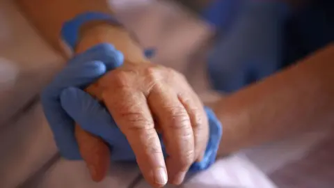 Getty Images A hand of a patient being held by a healthcare worker wearing a blue plastic glove. The patient has a blue wristband on.