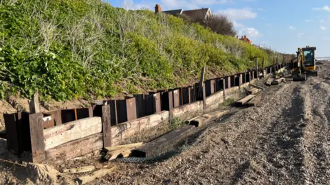 Darren Rozier/BBC Sheets of metal and wood being installed where the edge of the beach meets a large, inclined grass verge. A digger is on the beach on the right of the image.