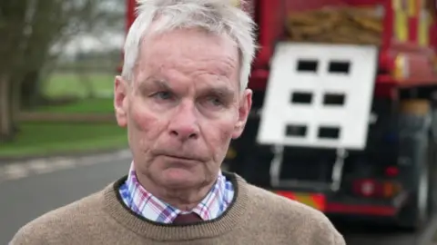 Martin has short grey hair and blue eyes. He is wearing a beige jumper with a brown border along the collar rim, over a blue and red check shirt. He is looking sternly towards the right of the camera. He is standing in a residential street with a green space behind him on the left and on the right is a fire engine in the distance, all of which is out of focus.