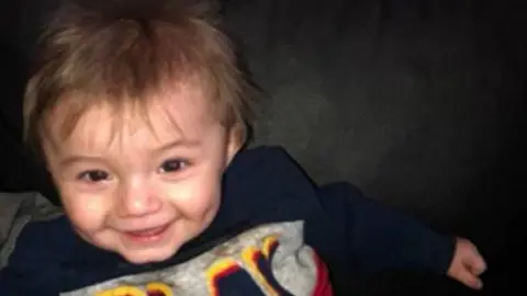 Ethan, a smiling young boy with dark hair, wearing a blue, grey and red jumper that says "Play" on it. 