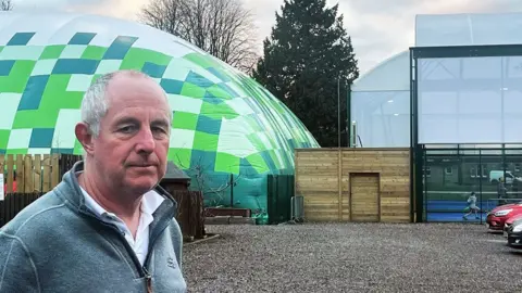 Keith Hughes Keith Hughes has low gray hair. He stands next to the green inflatable tennis court.