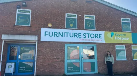 A woman in a green striped top and black trousers stands outside a red brick two-storey building with a white, green and blue sign saying Furniture Store, Severn Hospice. The windows are covered with adverts for electricals, homeware and furniture, and there are two double doors.