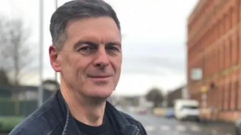 Shaun Harkin standing on a street with buildings, a road and cars behind him. He has black hair and wears a black jacket.