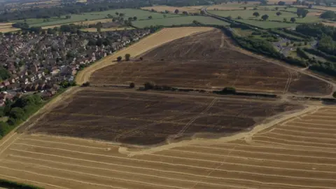 BBC/Mark Graham Large fields. A huge part of the fields are burnt and darkened. A road is to one side of the fields but houses are to the other.